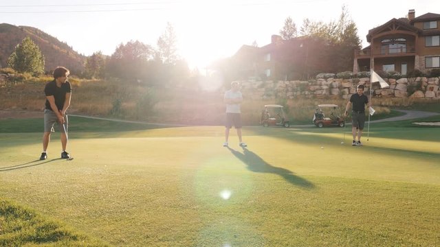 Camera Rises On The Green As Man Putts Golf Ball And Others Watch. The Camera Moves From A Very Low Angle Behind A Bunker And As It Rises, It Reveals 3 Men Playing Golf. One Is Hitting The Ball.