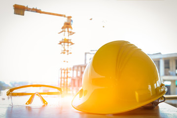 Construction safety concept, yellow hard safety helmet hat and harness protective gloves hard and yellow safety glasses on wooden with tower crane in background, spot fucus.