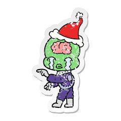 distressed sticker cartoon of a big brain alien crying and pointing wearing santa hat