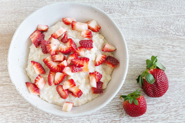 Top view of rolled  organic porridge with cut strawberries and two whole berries in marble bowl on wooden table  healthy breakfast