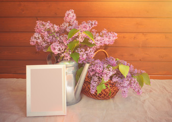 White frame,  watering can and basket with branches of lilac flower on table