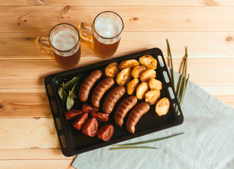 Two mugs of beer, griled pork  sausages, roasted potatoes and fresh tomatoes. Toned image.