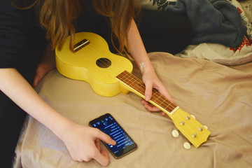 Teen girl tuning yellow soprano ukulele guitar by using application on smartphone while sitting in...