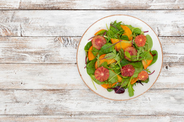 Salad (arugula, chard, mustard salad, spinach, persimmon, red orange, feta cheese, roasted pumpkin seeds, balsamic sauce cream) on white wooden background, top view