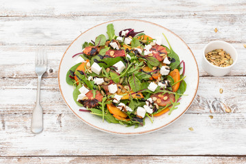 Salad (arugula, chard, mustard salad, spinach, persimmon, red orange, feta cheese, roasted pumpkin seeds, balsamic sauce cream) on white wooden background, top view