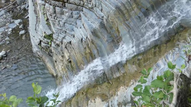 Small waterfall in the mountains, top view. Pure water flowing between stones and fall into the pool. Old rocks and plants at the sides, lot of foam in the stream. Sunny summer day, natural landscape.