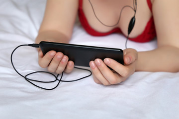 Girl in negligee lying on the bed with a smartphone in her hands. Sexy woman in headphones, concept of watching video, listening to music, social media, online communication, video chat