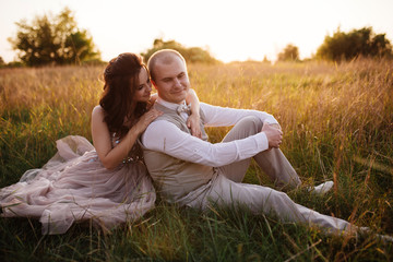 Wedding near the river in field at sunset with brown horse. Bride in light airy dress in color of dusty rose. Beige dress with sparkles. Light suit with bow tie. bride and groom embrace and kiss.