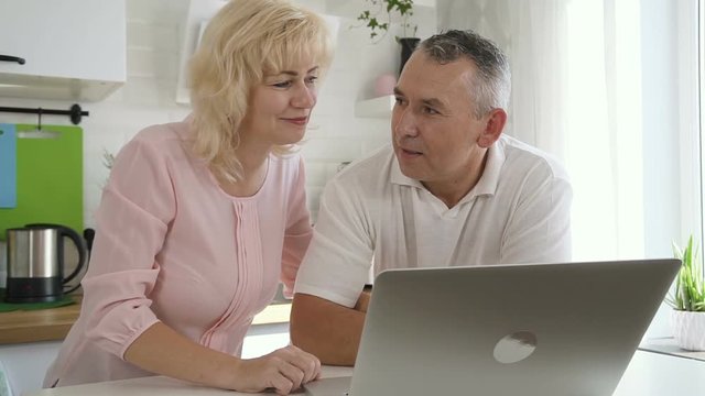 Happy elderly family using computer in apartment kitchen. Female pensioner and matre man standing near laptop and discussing.