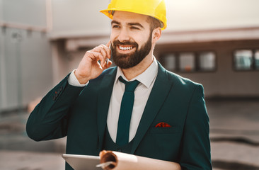 Portrait of bearded architect in formal wear talking on the phone and holding project and tablet while standing on construction site.