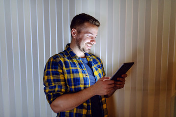 Smiling employee dressed in plaid shirt using tablet while leaning on wall in his office late at night. Side view.
