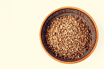 Dry raw buckwheat grains in brown ceramic bowl on white wooden background.