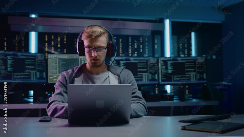 Poster Portrait of Software Developer / Hacker / Gamer Wearing Glasses and Headset Sitting at His Desk and Working / Playing on Laptop. In the Background Dark High Tech Environment with Multiple Displays.