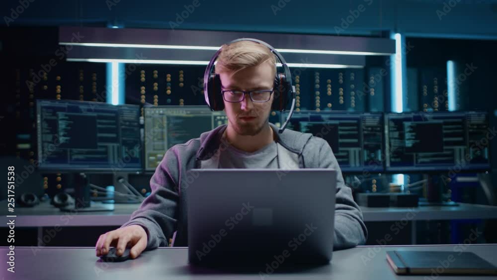 Aufkleber Portrait of Software Developer / Hacker / Gamer Wearing Glasses and Headset Sitting at His Desk and Working / Playing on Laptop. In the Background Dark High Tech Environment with Multiple Displays.