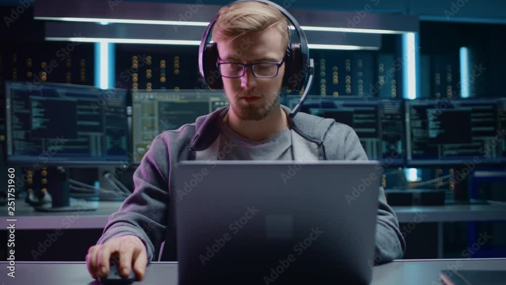 Aufkleber Portrait of Software Developer / Hacker / Gamer Wearing Glasses and Headset Sitting at His Desk and Working / Playing on Laptop. In the Background Dark High Tech Environment with Multiple Displays.