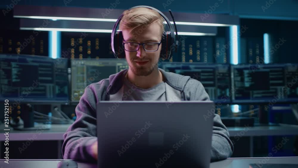 Aufkleber Portrait of Software Developer / Hacker / Gamer Wearing Glasses and Headset Sitting at His Desk and Working / Playing on Laptop. In the Background Dark High Tech Environment with Multiple Displays.