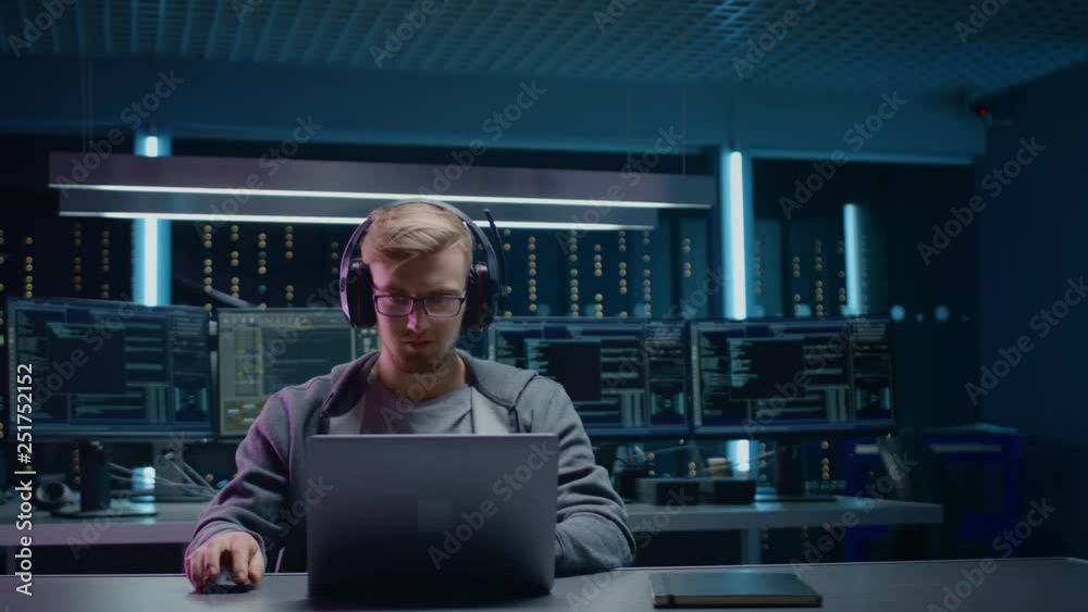 Poster Portrait of Software Developer / Hacker / Gamer Wearing Glasses and Headset Sitting at His Desk and Working / Playing on Laptop. In Background Dark Neon High Tech Environment with Multiple Displays.