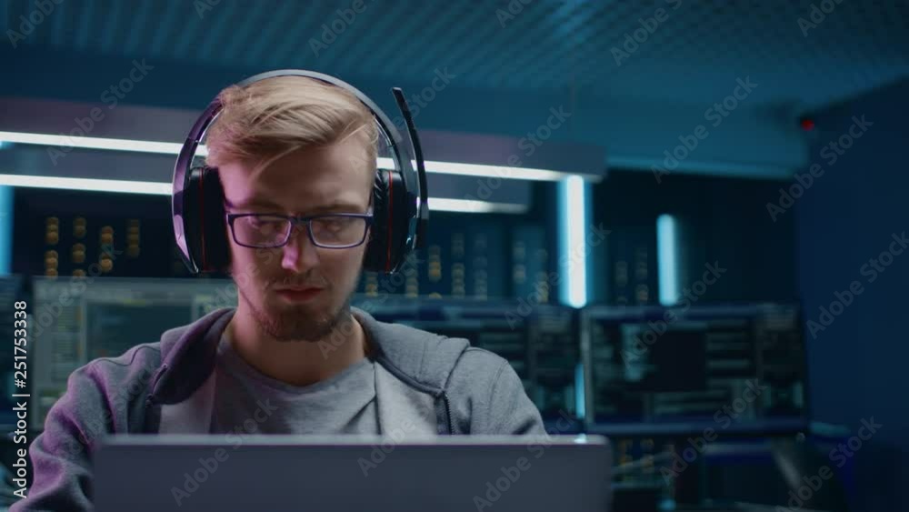 Poster Portrait of Software Developer / Hacker / Gamer Wearing Glasses and Headset Sitting at His Desk and Working / Playing on Laptop. In Background Dark Neon High Tech Environment with Multiple Displays