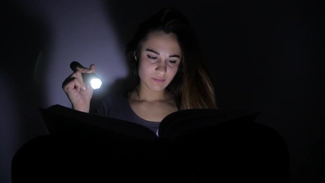 close-up portrait girl is reading a book with a flashlight in a dark room at night. 4k. Motion shooting