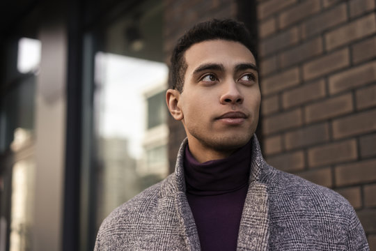 Closeup portrait of handsome African American man with positive face waiting for taxi outdoors. Successful fashion model wearing stylish grey coat and purple turtleneck walking along urban street