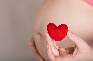 Obraz premium Young pregnant woman keeps small red plush heart close to her belly.