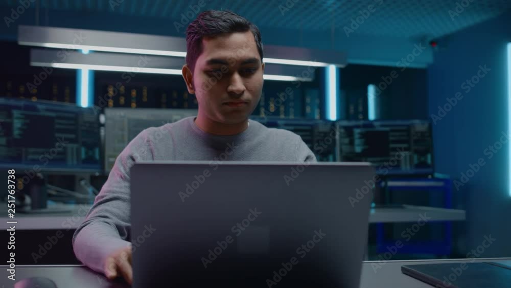 Poster Portrait of Software Developer / Hacker / Programmer Sitting at His Desk and Working on Laptop Computer In the Background Dark Neon High Tech Environment with Multiple Displays. Zoom Out Shot