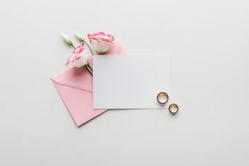 top view of empty card with pink envelope, flowers and golden wedding rings on grey background