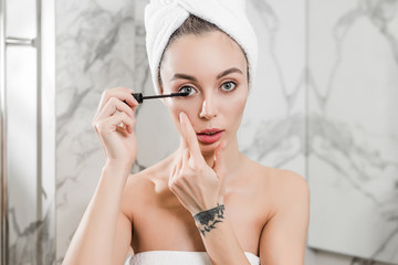 Young woman applying black mascara on eyelashes with brush wrapped in towels in the bathroom.  beauty makeup and skincare concept