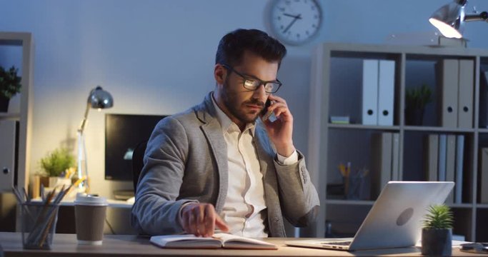 Handsome Caucasian young man in glasses working at night on the laptop in the office, calling somebody on the mobile phone and talking about the business.
