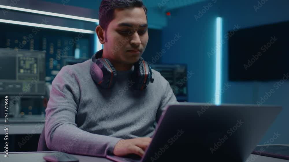 Aufkleber Portrait of Software Developer / Hacker / Programmer Sitting at His Desk and Working on Laptop Computer In the Background Dark Neon High Tech Environment with Multiple Displays. 