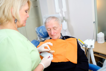 Obraz premium Senior man 70-75 years old in dentist chair, check and select color of teeth. Dentist makes the process of treatment in dental clinic. Dentistry, medicine and health care concept
