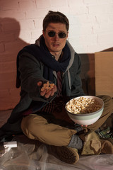 Fototapeta na wymiar homeless man holding popcorn in raised hand while sitting by white brick wall
