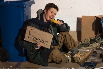 homeless man eating corn cob and holding cardboard card with "freegan" inscription