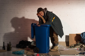 homeless man holding bread baguette near face while leaning on trash container