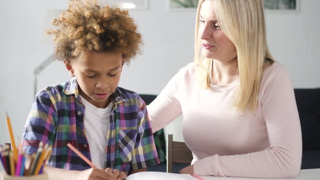 American adult woman is teaching and helping preteen hispanic boy with writing homework at the desk. Indoors studying with female tutor together. Happy school time and family love to education process