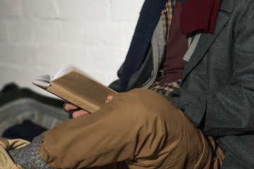 cropped view of homeless man reading book while sitting by white brick wall