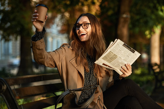 Photo of happy woman drinking takeaway coffee and reading newspaper while sitting on bench in sunlit alley