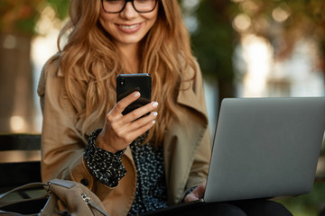 Photo of stylish girl using mobile phone and laptop while sitting on bench in sunlit alley