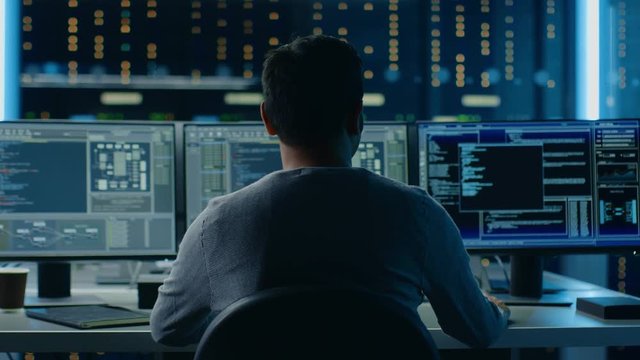 IT Specialist Working on Personal Computer with Monitors Showing Coding Language Program with System Monitoring Interface. In the Background Technical Room of Data Center. Back to the Camera Shot