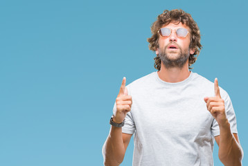 Handsome hispanic model man wearing sunglasses over isolated background amazed and surprised looking up and pointing with fingers and raised arms.