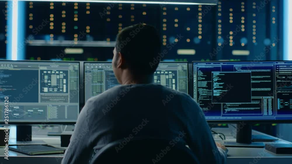 Poster IT Specialist Takes His Place at the Desk and Starts Working on Personal Computer with Screens Showing Software Program with Coding Language Interface. In the Background Technical Room of Data Center.