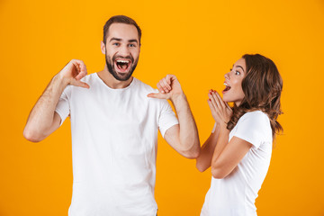 Portrait of caucasian couple man and woman in basic clothing pointing fingers, while standing together isolated over yellow background