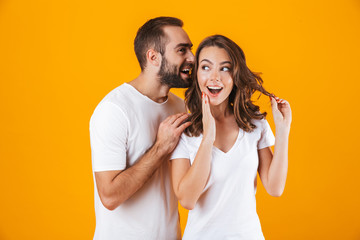 Image of attractive man whispering secret or interesting gossip to woman in her ear, isolated over yellow background