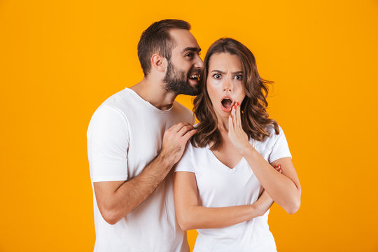 Image of content man whispering secret or interesting gossip to woman in her ear, isolated over yellow background
