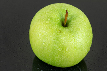 A fresh green apple with water droplets on its surface, placed on a sleek black reflective surface, showcasing its vibrant color and freshness.
