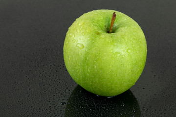A fresh green apple with water droplets on its surface, placed on a sleek black reflective surface, showcasing its vibrant color and freshness.