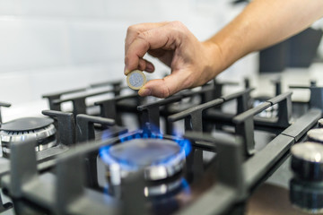 Cooktop with burning gas ring with hands holding coin of 1 euro for combustion at home.