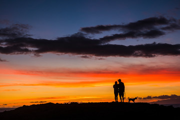 Fototapeta premium Romantic couple wioth dog enjoying and looking the sunset with red and orange coloured clouds in the sky - wanderlust and travel concept for happy people - dawn and colors background concept