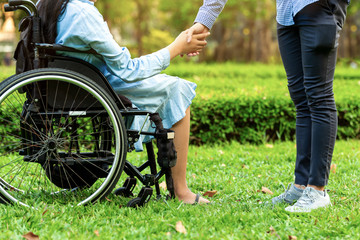 Young woman in wheelchair holding hands with caretaker man in public park.