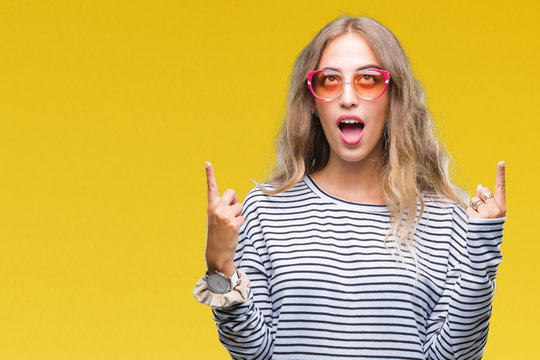 Beautiful young blonde woman wearing sunglasses over isolated background amazed and surprised looking up and pointing with fingers and raised arms.